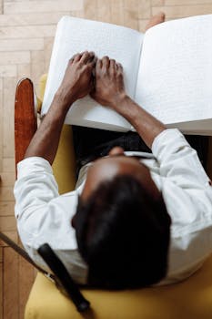 A blind man reading a Braille book indoors, focusing on touch and concentration.